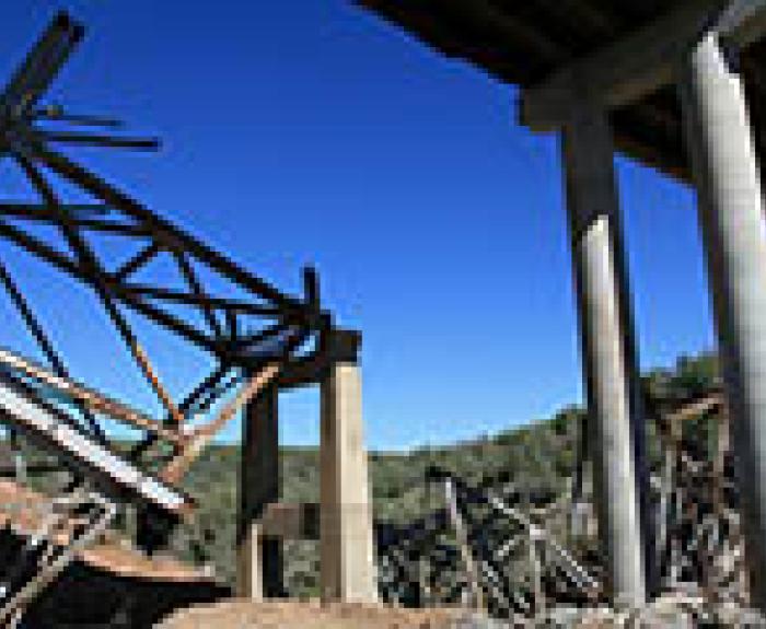 Collapsed bridge with twisted metal beams and concrete pillars under a clear blue sky.