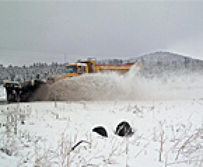 A snow plow clears snow from a road in a snowy landscape, spraying snow to the side. Snow-covered grass and hills are visible in the background under a cloudy sky.