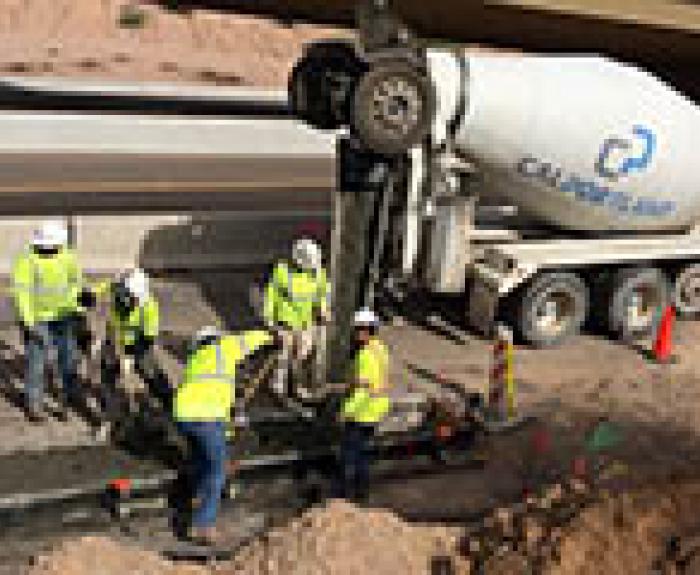 Construction workers in safety vests pour and level concrete from a cement truck at a roadside construction site.