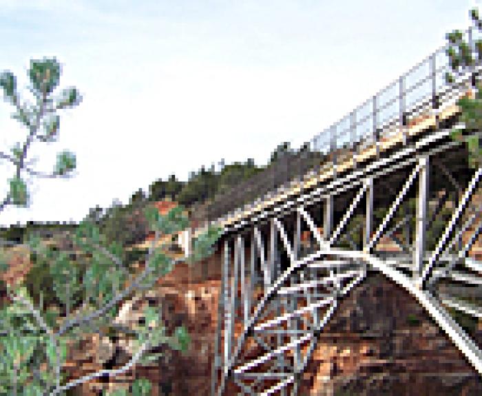 Steel arch Midgley Bridge in northern Arizona spans a rocky canyon with trees and greenery in the foreground under a clear sky.