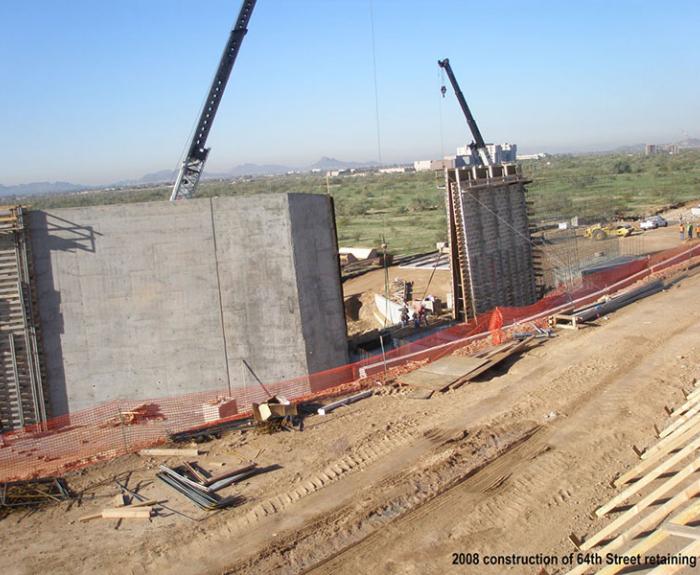 Construction site with cranes and workers building a large retaining wall, surrounded by dirt, materials, and safety barriers.