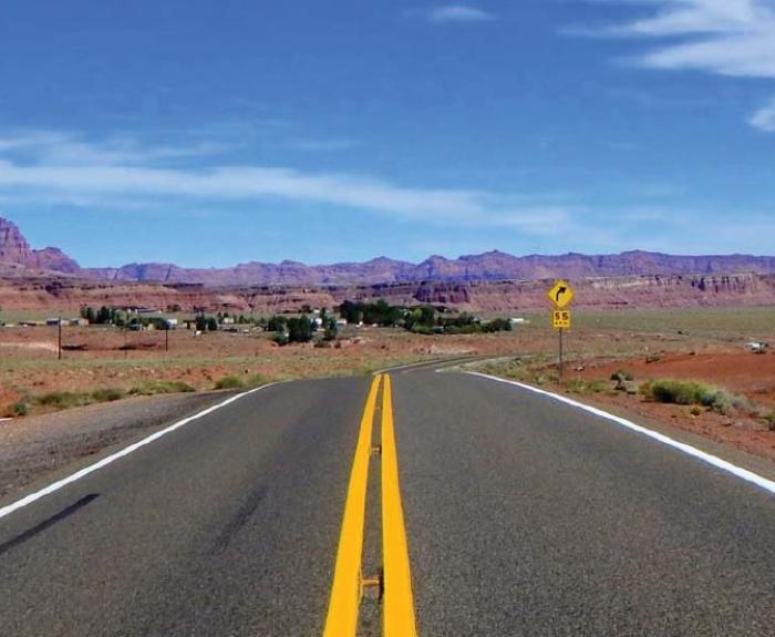 A two-lane highway with a double yellow line runs through a desert landscape with red rock formations under a blue sky. A road sign and small buildings are visible in the distance.