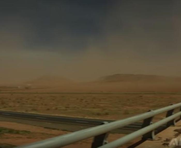 Dust storm approaching a desert highway, with guardrails and distant hills under a dark sky.