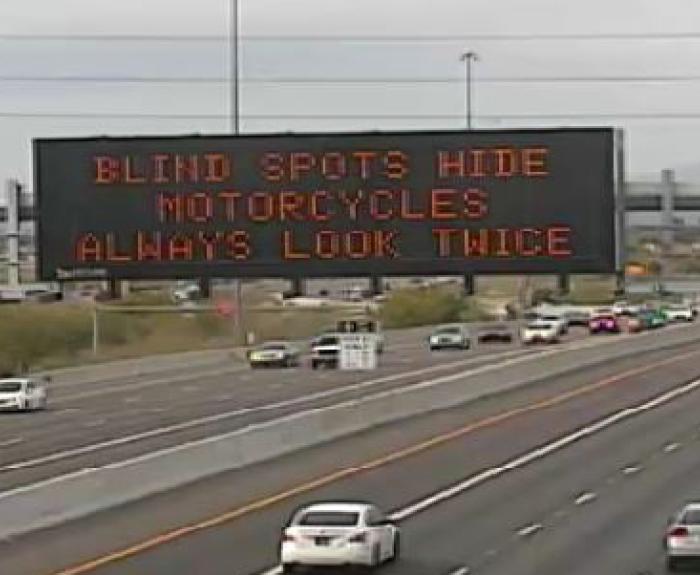 Highway electronic message board reads BLIND SPOTS HIDE MOTORCYCLES ALWAYS LOOK TWICE above multiple lanes of traffic.