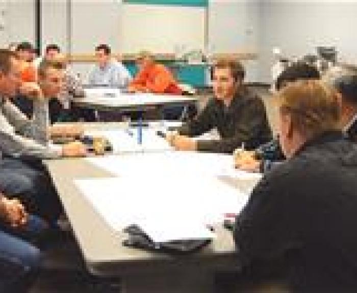 A group of people sit around tables in a meeting room, discussing and writing on large sheets of paper.
