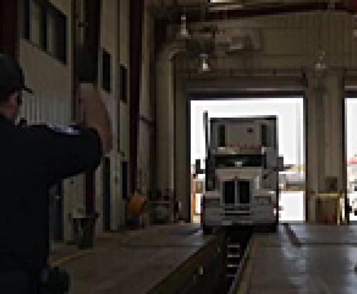 An officer signals a white semi-truck to enter an inspection bay.