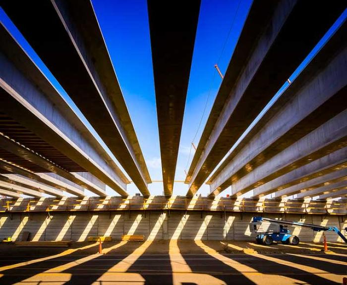 View from beneath a bridge under construction, showing parallel concrete beams and a blue sky above, with a construction lift vehicle on the right.