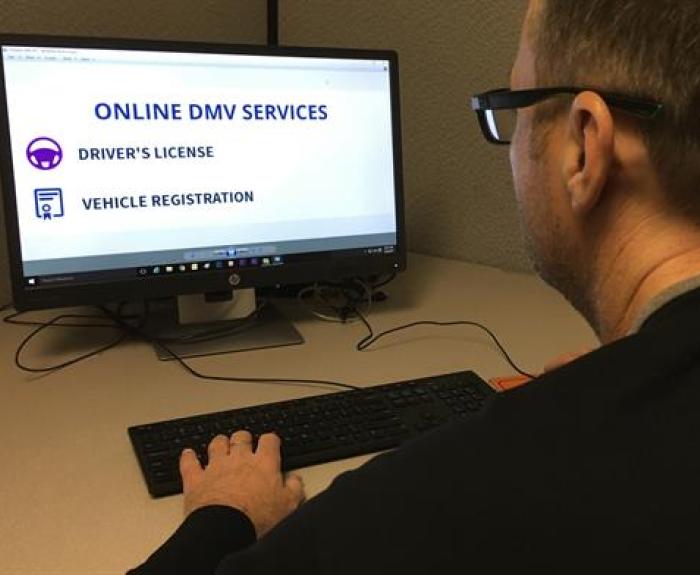 A man sits at a desk using a computer displaying online DMV services, with options for drivers license and vehicle registration.