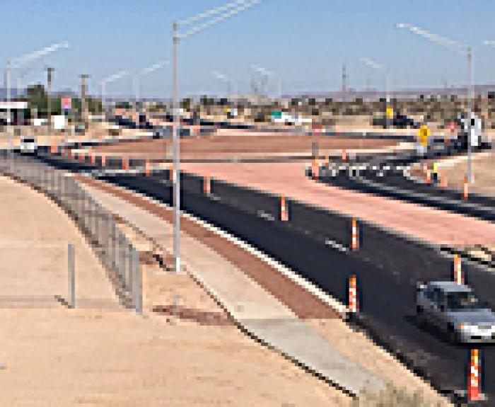 A newly paved multi-lane road with traffic cones and construction signage, surrounded by a dry, desert landscape under a clear sky.
