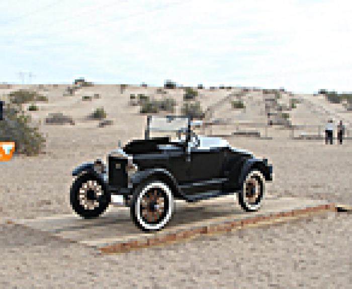 A vintage black car is displayed on a wooden platform in a sandy desert area with a sign nearby.