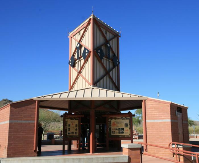 A brick and metal visitor center with a tall tower structure, information boards, and a clear blue sky in the background.