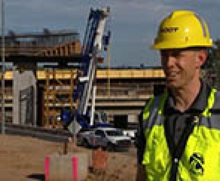 A construction worker in a yellow safety vest and hard hat stands at a building site with cranes, vehicles, and an overpass structure in the background.