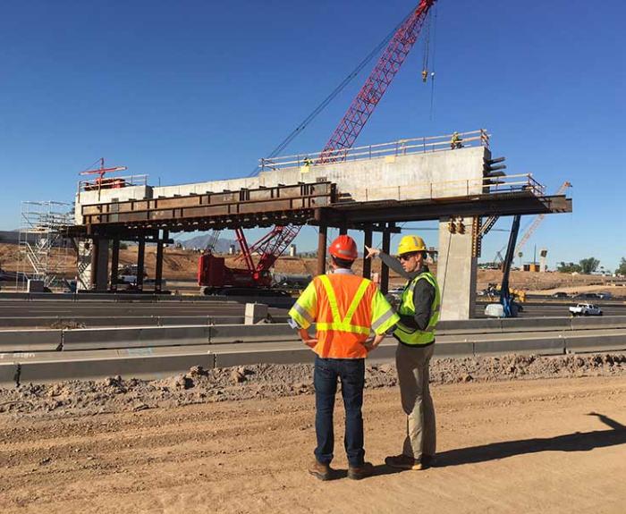 Two construction workers in safety gear stand on a dirt road, observing a partially built bridge with cranes and equipment on a clear day.