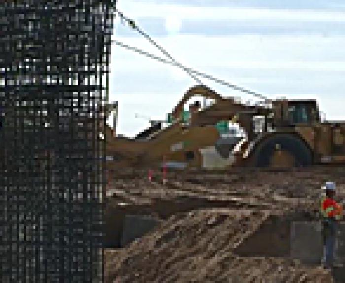 Construction workers in safety gear stand near a steel rebar structure, with heavy machinery operating on a dirt site in the background.
