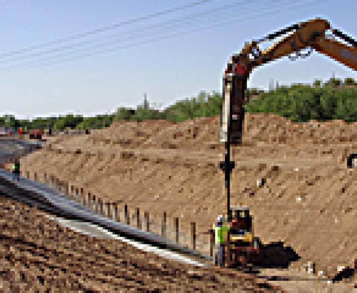 Workers and an excavator build a canal or ditch, lining it with material on a sunny day.