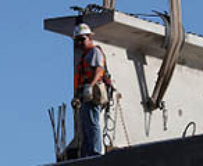 Construction worker in safety gear stands on a beam near large concrete structure under blue sky.