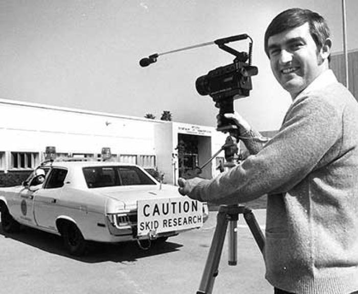Black and white image of a man with a camera standing in front of a car that has a "Caution: Skid Research" sign on the bumper