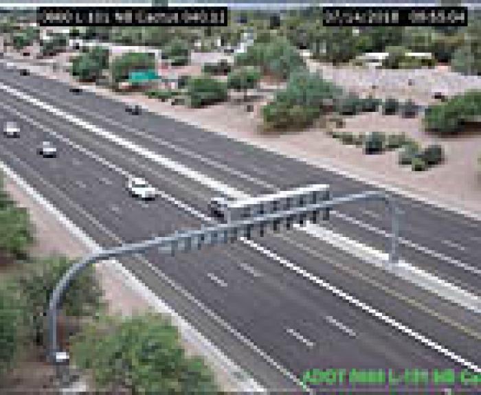 A highway with several vehicles traveling in both directions, surrounded by desert landscaping and trees, viewed from an overhead traffic camera.