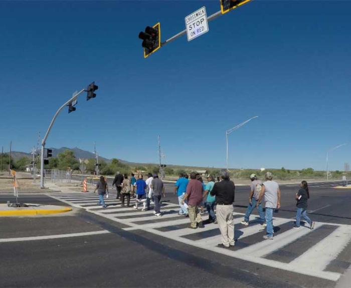 A group of people crosses a wide street at a crosswalk under a clear blue sky and traffic lights.
