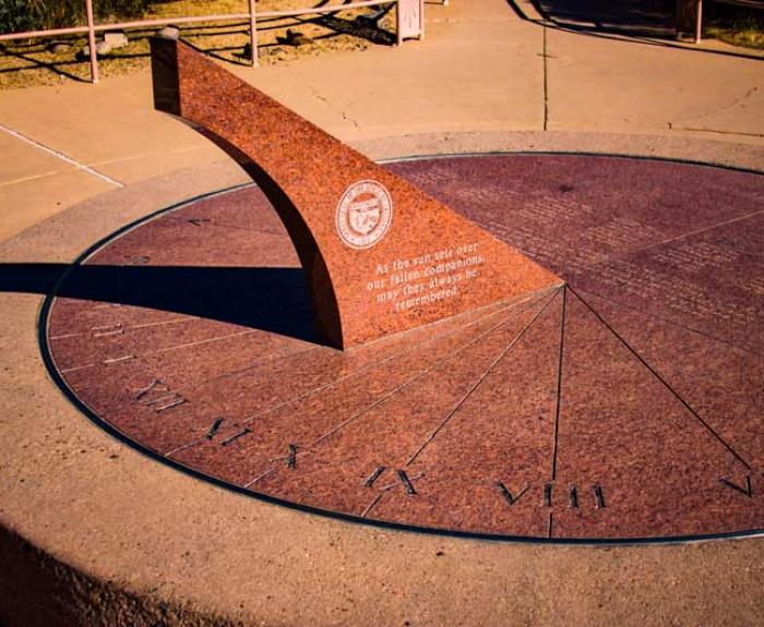 Large stone sundial with Roman numerals, an upright triangular gnomon, and an inscribed text in the center, set in a paved outdoor area.