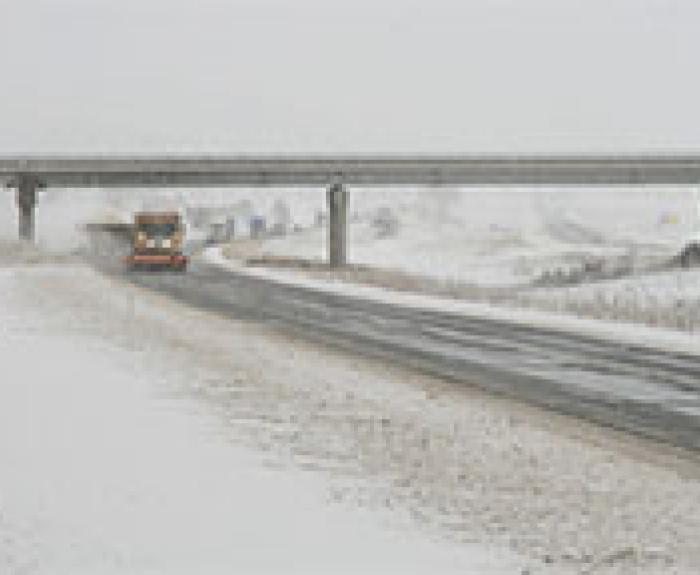 A snowplow clears a snow-covered road beneath an overpass on a wintry day.