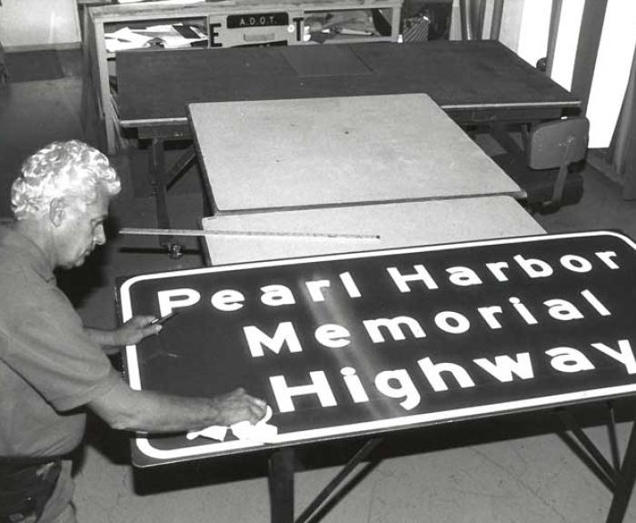 Black and white photo of a man working on a Pearl Harbor Memorial Highway sign.