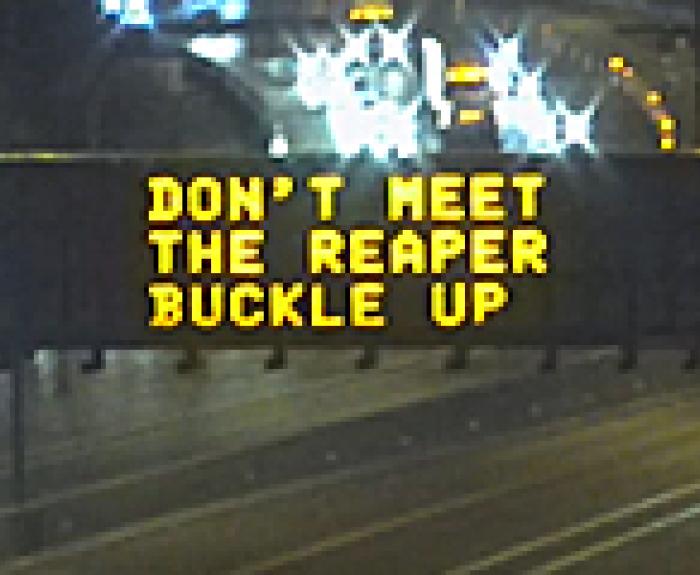 Highway electronic sign at night displays the message: DON’T MEET THE REAPER BUCKLE UP above lanes of traffic.