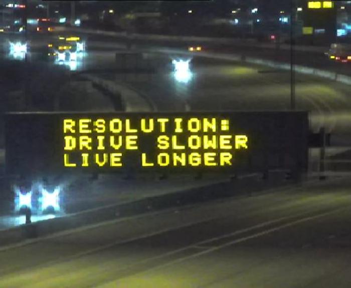 An electronic highway sign displays the message: RESOLUTION: DRIVE SLOWER LIVE LONGER to encourage safe driving at night.