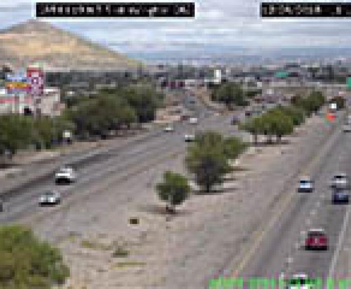 Wide view of a multi-lane highway with moderate traffic, bordered by trees and buildings, with a hill in the background under a cloudy sky.