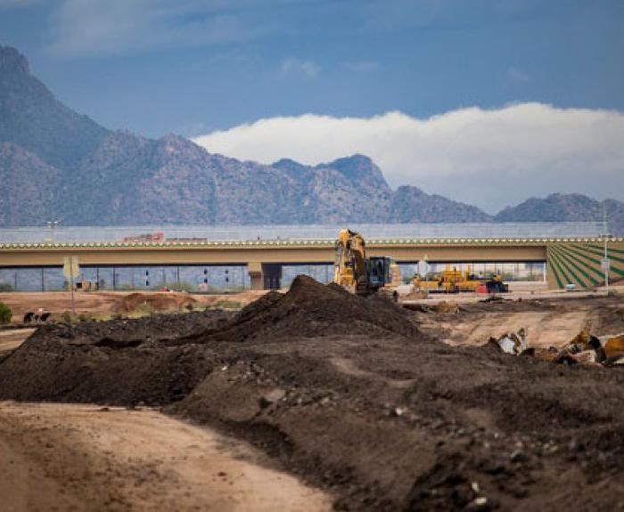 Excavators work on a dirt construction site near a bridge with mountains in the background.
