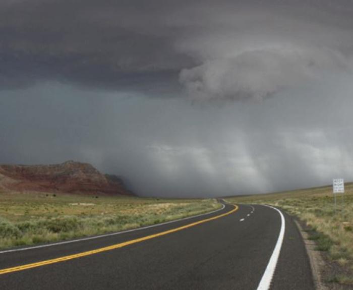 A winding road leads to distant mountains under a dark, stormy sky with rain falling in the distance.
