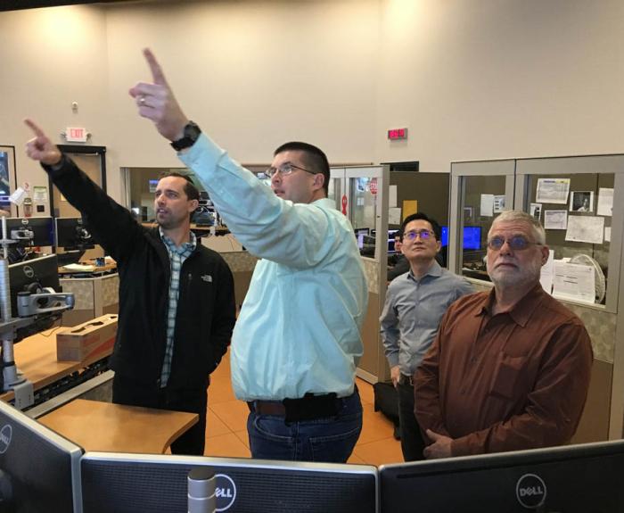 Four men stand in an office, two of them pointing at monitors, while the others observe. Computer screens and office equipment are visible.