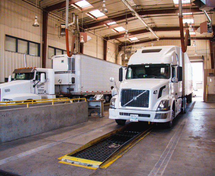Two white semi-trucks inside a large ADOT inspection facility with high ceilings and overhead lights.