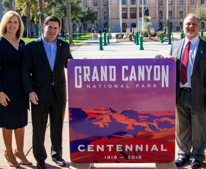 Three people stand outdoors holding a Grand Canyon National Park Centennial sign, 1919-2019.