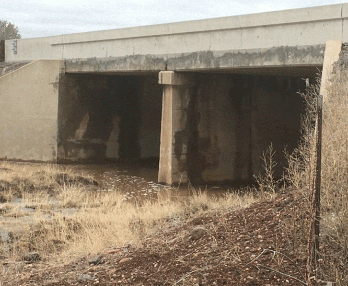Concrete bridge over a shallow stream with dry grass and rocks in the foreground; stained walls indicate water flow levels.