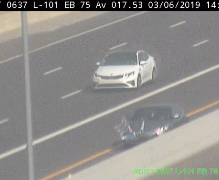 Traffic camera view of two cars traveling on a multi-lane highway; one white car in the lane, one darker car with flags on the shoulder. A ladder appears to be caught on the dark car's hood.