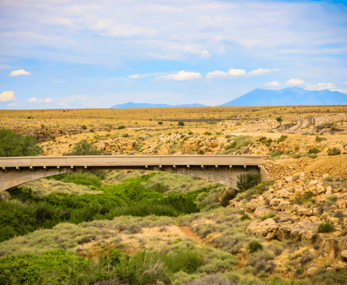 A concrete bridge spans a green, brush-filled arroyo in a dry desert landscape with distant mountains and a partly cloudy sky in the background.