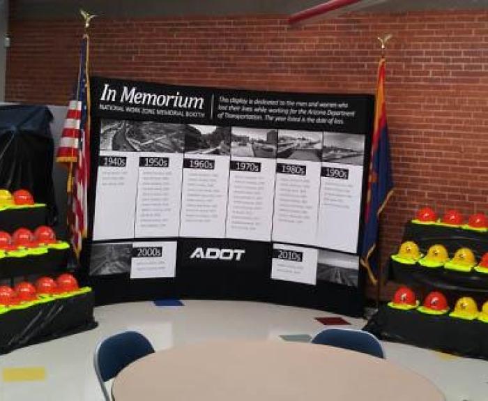 A display of orange and yellow hard hats honors fallen highway workers, with a memorial banner and US flags in the background.