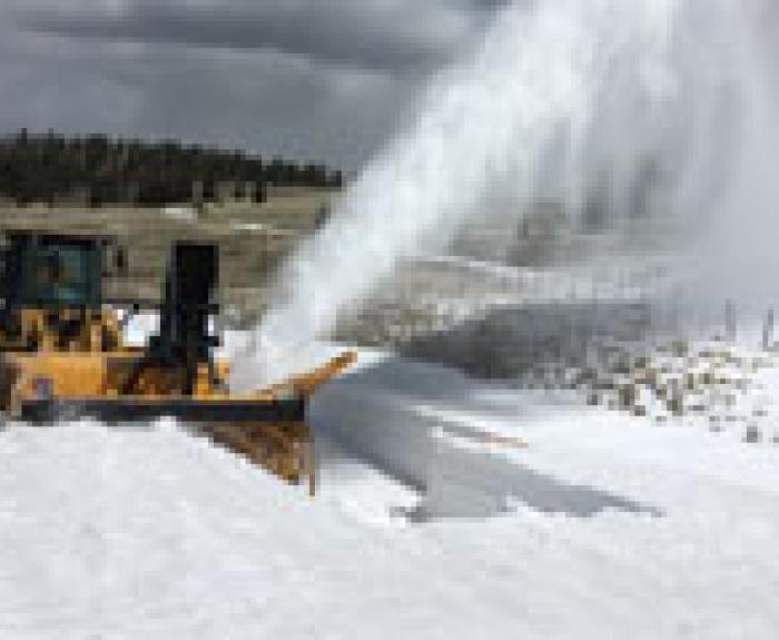 A snowplow clears snow from a road, blowing it to the side, with cars stopped on the roadway and a grassy field in the background.