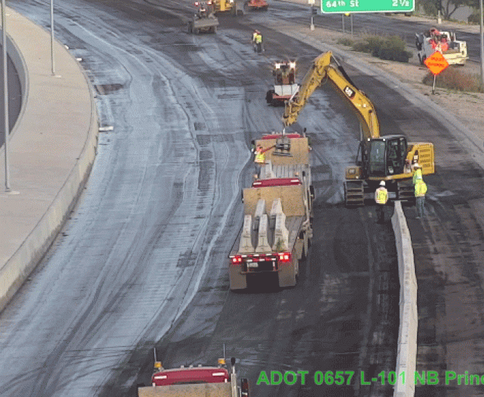 Construction workers and machinery lay concrete barriers on a multi-lane highway with traffic signs and equipment visible along the road.