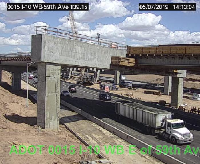 A white semi-truck drives under a concrete overpass under construction, with cranes and unfinished sections visible; blue sky with scattered clouds above.