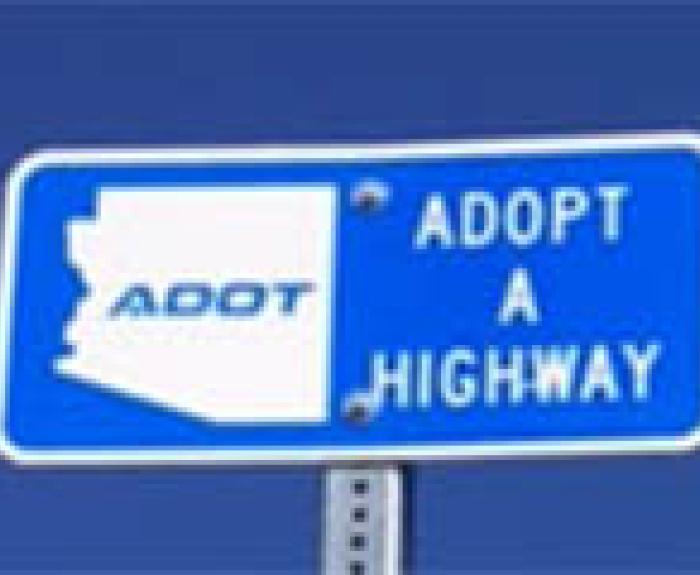 Blue road sign reads ADOT Adopt A Highway with an outline of the state of Arizona and the letters ADOT inside the outline, against a clear blue sky.