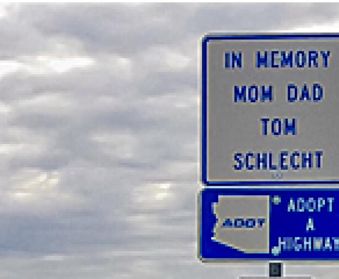 A roadside sign reads In Memory Mom Dad Tom Schlecht above a sign showing the outline of Arizona and text ADOT Adopt a Highway. Cloudy sky in the background.