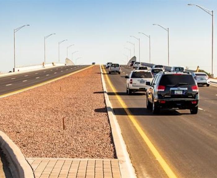 Several vehicles drive up a divided highway overpass on a clear day, with streetlights lining both sides and a rocky median in the center.