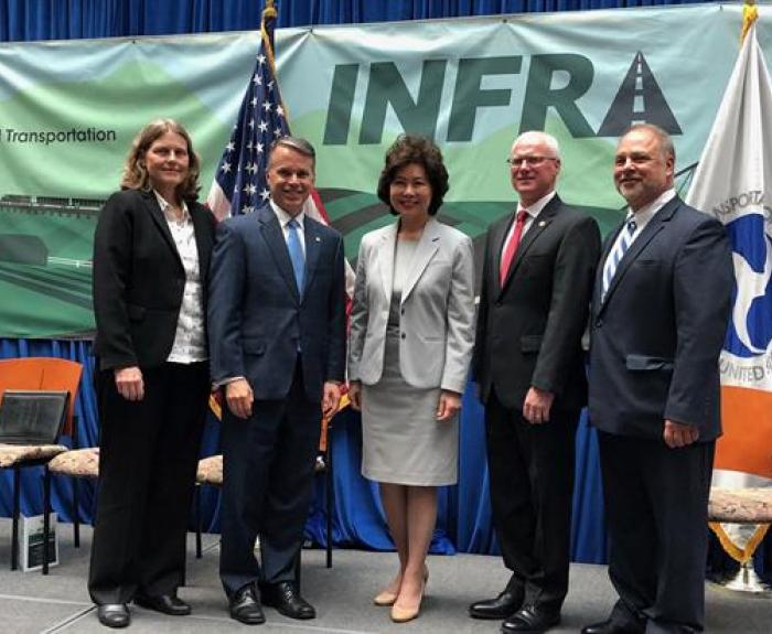 Five people in business attire pose for a group photo in front of a transportation-themed banner and U.S. flags at an official event.