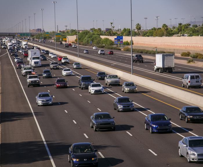 Multiple lanes of vehicles traveling in both directions on a busy highway bordered by concrete barriers and roadside signs under clear skies.