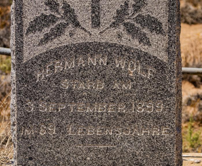 A weathered gravestone curved at the top with a cross and leaves carved into the stone. The name Hermann Wolf is carved into the stone along with birth and death dates.