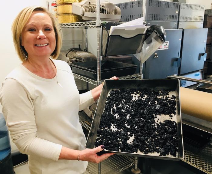 A woman stands in a laboratory holding a metal tray filled with dried black material, with shelves and lab equipment in the background.