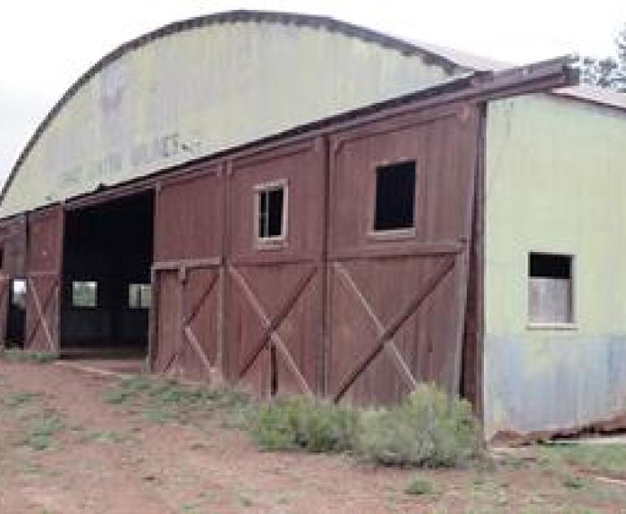 A large, weathered barn with wooden doors and windows, featuring a partially open entrance and faded green metal siding. Shrubs and dirt surround the structure.