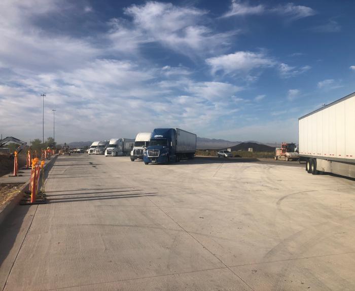 Several semi-trucks parked on a wide concrete lot under a partly cloudy sky with mountains in the distance.